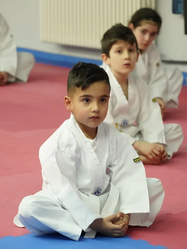 Kinder im weißen Taekwondo-Anzug sitzen aufmerksam auf der Trainingsmatte.
